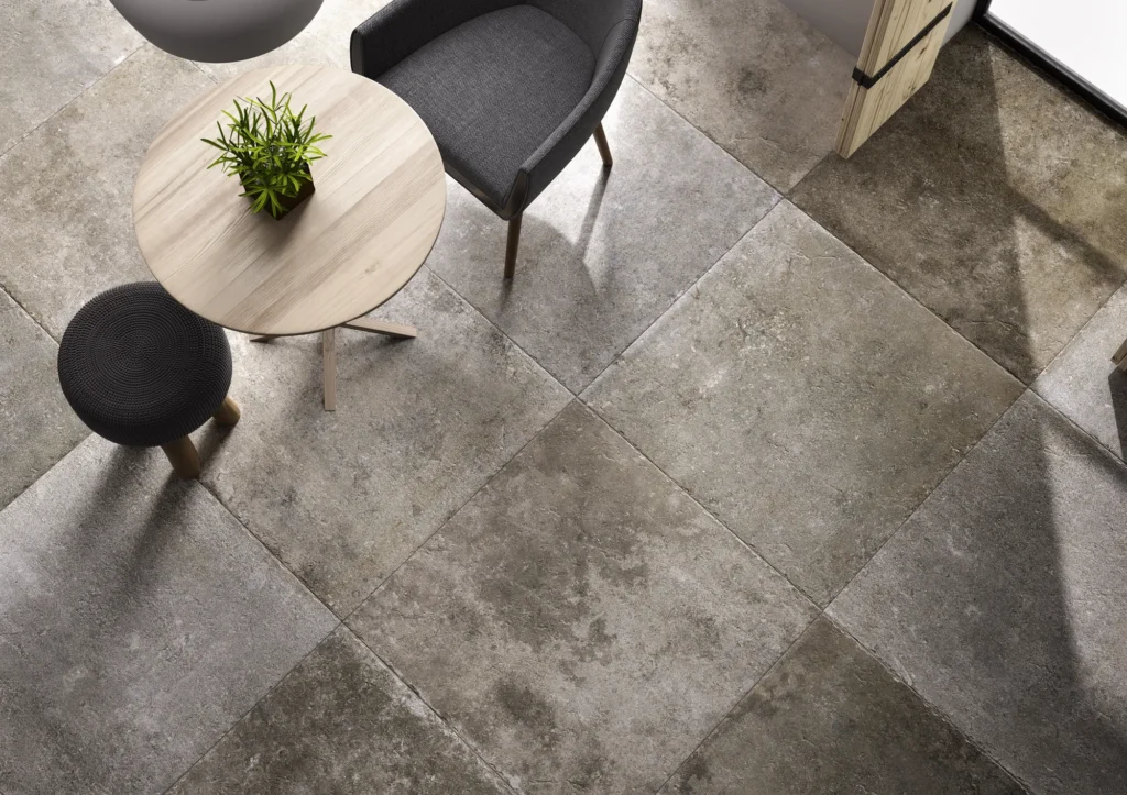 Overhead view of a modern room with large gray stone tiles. Features a round wooden table, potted plant, gray chair, and a matching gray stool. Minimalist and serene.